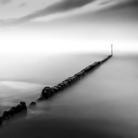 A black and white image of groynes in the sea leading into white haze. A triangular sign marks the end of the groyne. Due to the long exposure, the sea is smooth. Cloud streaks can be seen in the sky.