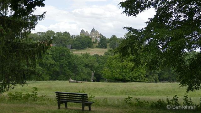 Im Vordergrund eine leere Sitzbank, dahinter Grasland und ein kleines Wäldchen. Auf einem Berg am Horizont die Sababurg. Das Bild ist rechts und links durch Bäume begrenzt.