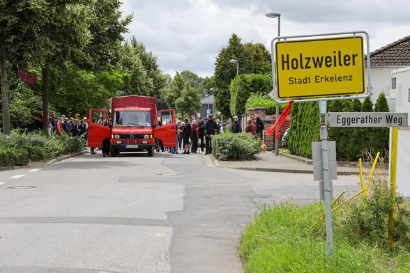 Der Blick von der L12 vorbei am Ortsschild vom Holzweiler auf den Dorfspaziergang, der vom Bündnismobil angeführt wird