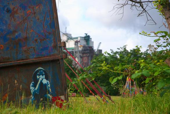auf einer Metalltür ist ein kleines hockende kind mit kapuzenpulli gesprüht.. im hintergrund eine wiese, dahinter ein riesieges industriewerk