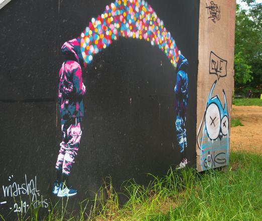 auf einer dunklen sperrholzwand sind zwei personen in stencilstile gesprüht, beide personen haben eine kapuzenjacke an und aus denen fliegen jeweil bunte kugeln in einander