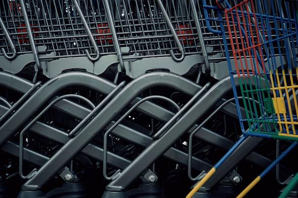 Side view of a row of metal-grey shopping carts in storage. Neither the wheels nor the gandles of the carts are in frame, they‘re just repeating metal structures. On the right-hand side there‘s a colourful children‘s shopping cart partly visible, rougly the rear quarter is in frame.