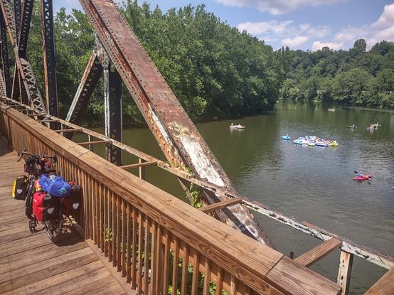 An old bridge turned into a scenic overlook over the Yough River with a loaded touring bike on it. There are kayaker and rafters floating in the river.