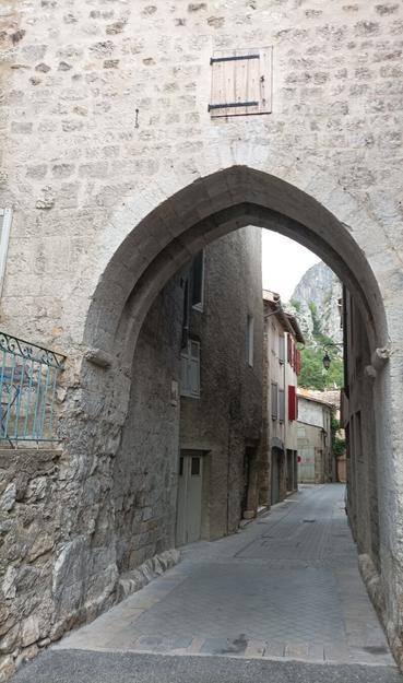 Stone-made arch, leading onto a narrow road with other stone made buildings. A high rock at the back end.