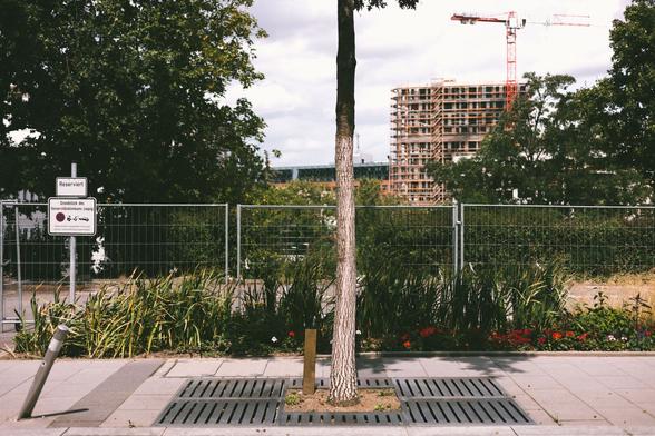 Ein Gehweg mit einer Baumplatte. Ein umgefahrener Poller und im Hintergrund ein Baukran an einem unfertigen Hochhaus. Zwischen all dem Beton