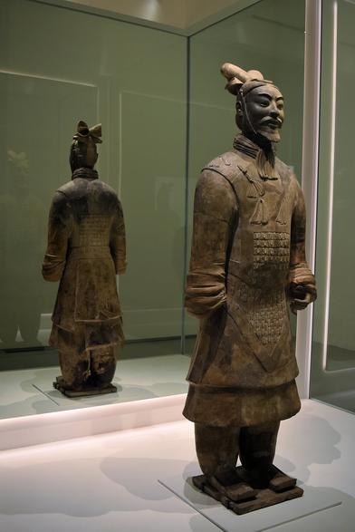 Large terracotta figure in the form of a Chinese army general, wearing a long, tunic-like uniform and baggy trousers. The figures is reflected in a mirror behind, which shows the view from the back