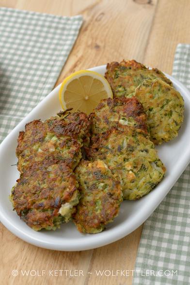 Photograph shows a plate of eight Kolokithokeftedes - Greek courgette cakes/patties - and half a lemon, arranged on a wooden dining table. On both sides of the plate the edges of green and white chequered placemats are visible.
