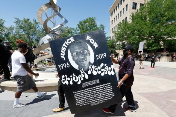 Photo taken during the 2020 protests, demanding justice in the death of Elijah McClain. Activists carrying signs march in the streets. In the foreground two protesters are carrying a huge sign with a picture of Elisha and text that reads:
"JUSTICE FOR ELIJAH MCCLAIN – 1996-2019" at the bottom, above his final words appears text reading: "@JusticeForElijahMcClain".