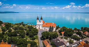 Uitzicht op het Balatonmeer vanaf de kerk in Tihany. De kerk staat in het midden. Het is een wit gebouw met twee torentjes en een oranje dak. Verder zijn er veel bomen en op de achtergrond het blauw-groene water van het meer.