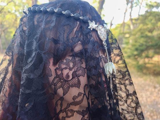Photo of a white woman under a lace veil. On her head she wears a string of prayer beads made from volcanic rock, rattlesnake vertebrae, a silver talisman, silver chain, a silver flower, and a steel key.