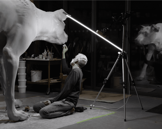 Photographie de Ron Mueck dans son atelier. 
A genou, portant un masque de protection, l'artiste travaille sur une immense sculpture de chien.
