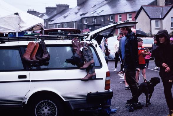 A colour film photograph of farmers market in Skibbereen, co. Cork, Ireland. There is an old white Volvo estate with pairs of cowboy boots hanging from the roof rack. A man with a black boxer dog is looking into the open booth of the car.