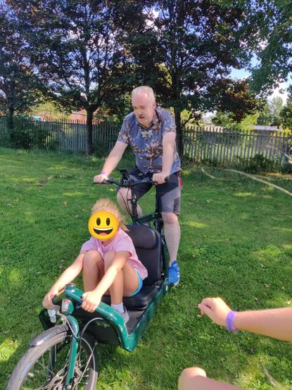Me riding a green cargo bike in a grassy field.  In the "cargo" section of the bike there's a seat and a child getting a ride.