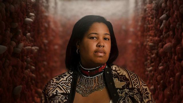 A young Black, Indigenous woman whose straight black hair is loosely tied back looks sideways at the camera. An earring and a nose ring  gleam in the overhead light, and she wears a tan-and-white jacket with plant, cloud, and geometric patterns. On her neck is an embroidered collar with a row of metallic medallions on chains draped from the front edge, covering her chest.