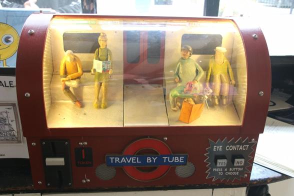 A model of part of an underground carriage. Four carved figures are sitting on seats under the windows. A carved child figure is peering into a shopping bag on the floor. The central door area is empty.