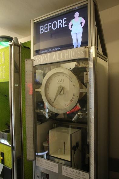 A large machine with a central "BMI" dial. Above is a screen showing a picture of a plump Angela Merkel and captioned "before".