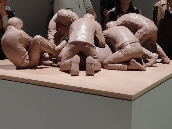 Photographie d'une petite sculpture en argile exposée sur un socle. 
Cinq hommes, penchés ou à genoux, semblent être en train de contraindre quelqu'un allongé au sol. 
Ils exercent des pressions sur son corps, voire des étirements. L'un des hommes a un pied posé sur ce corps. 
Sur le personnage de droite, on distingue l'empreinte laissée par les doigts du sculpteur Ron Mueck.