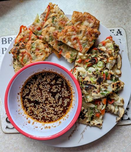 Slices of vegetable pancakes beside a small bowl with a dark sauce that is topped with sesame seeds.