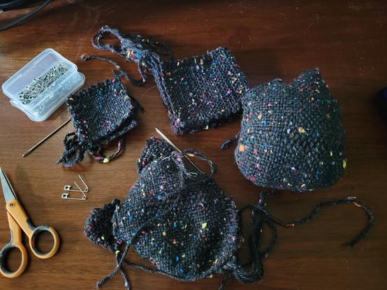 Photo is of handwoven squares of yarn in black with colored speckles, oranfe handled scissors, needle and safety pins against a wood surface.
