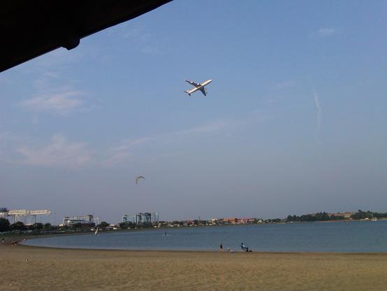 Looking across the beach, at water's edge, a couple of people have a chair and blanket set up. Across the bay we can see the metal structures over the rail yard. Castle Island is visible in the distance. A person in  the water is pulled by  a parasail above. Higher up in the sky, a jet ascends from Logan Airport.