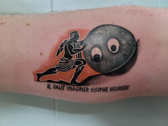 A tattoo depicting the Greek myth of Sisyphus pushing a boulder up a hill. The boulder in this case is rendered in a realistic detail with two googly eyes on its center as reference to the movie Everything Everywhere All At Once. Below the image is the text “I imagine Sisyphus to be happy” in French, as concluded by the 1942 philosophical essay by Albert Camus