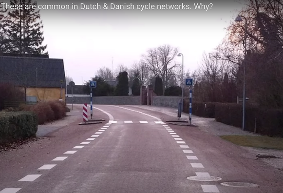 picture from ~20m in the video, a chokepoint with curbs and posts on each side of the road, allowing only one direction of car travel to go through the crosswalk at a time, shortening the exposed crossing distance, and giving people on bikes a way around the outside