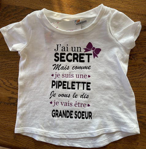 T-shirt enfant blanc pour annoncer une naissance. Texte : j’ai un secret, mais comme je suis une pipelette je vous le dis je vais être grande sœur. Écriture noire et violette. Petit nœud papillon en haut à droite. ￼