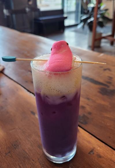 A toll narrow glass with bright purple liquid in it with foam on top and garnished with a pink marshmallow peep