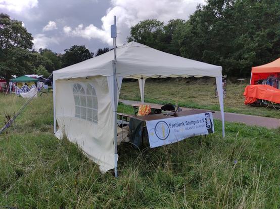 Info-Pavillon mit Biertischen und Freifunk Stuttgart e.V. Banner das an einem Biertisch befestigt ist. Eine Stange ragt vorne rechts nach oben. Am der Spitze ist ein Richtfunk-Gerät befestigt.