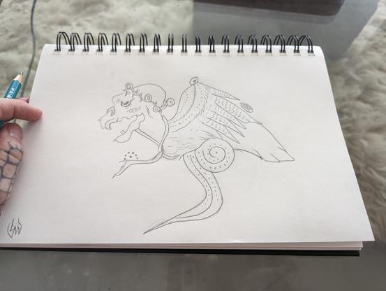 A sketchbook lays open, landscape oriented, on a glass table through which a white wool rug is visible. A hand lays loosely on the left side of the page holding a blue prismacolor pencil. On the page is a Mayan death owl, redrawn from a classic Mayan pot. It has a harpy-like head with a crown of stylized eyeballs and thorns, drifting tendrils ending in interesting dotwork, and connected wings that terminate in a thick feathered tail.