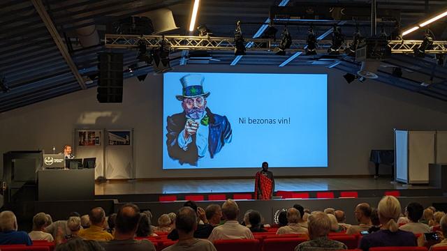 Parolanto en afrikanaj vestaĵoj staras antaŭ la publiko. La lumbildo montras la tekston "Ni bezonas vin!"