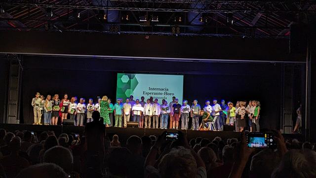 Homoj staras ĉe la prezentejo. Ili kantas en direkto de publiko. La lumbildo montras la tekston "Internacia Esperanto Ĥoro - Tutmonda".