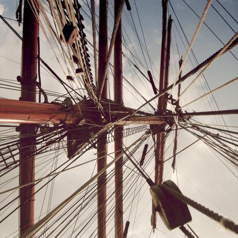 A worm‘s eye view of a sailing ship‘s mast, with a blue and lightly clouded sky in the background.