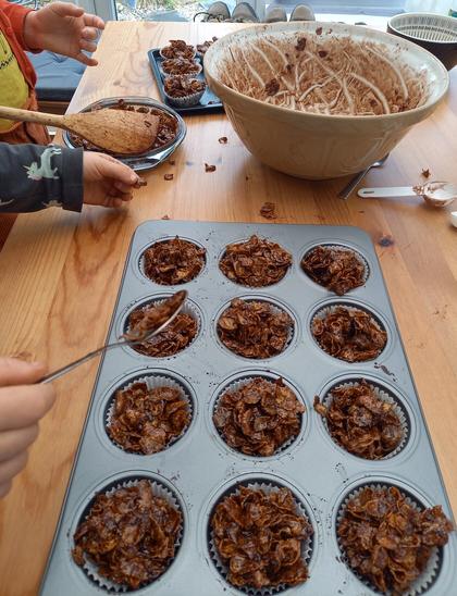 Children (just out of shot) making Cornflake Crispy Cakes. There's a tray of cakes and a chocolatey bowl with only a few stray cornflakes on the table.