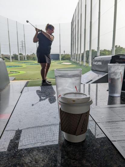 In the foreground, a cup of coffee and a water on a table. In the background, my wife in t, hitting a golf ball at Top Golf, a gamified driving range.