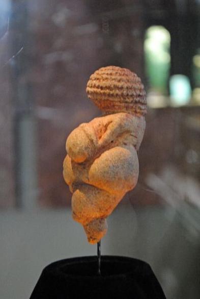 Photo of the Venus of Willendorf, mounted in a museum case. It's an ancient terracotta figurine of a fat lady, probably the most famous one- she's pear-shaped with a curly hairdo.
