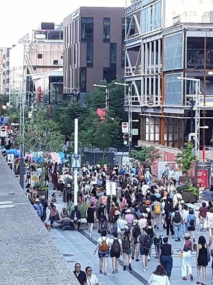 Manifestation pour le droit des personnes trans et non-binaires. Photo floue et zoomée à l'excès sur laquelle on voit des gens marcher vers l'Est sur la rue Sainte-Catherine à Montréal, à la hauteur du Quartier des spectacles. En arrière-plan, un énorme drapeau de la fierté trans tenu par une quinzaine de personnes.
