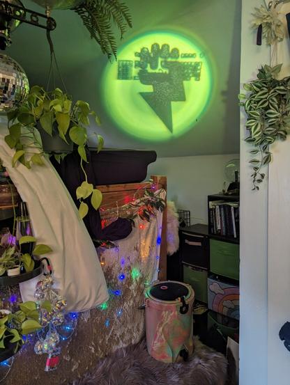 A picture of a room with a green circle projected on the wall. Inside the circle is an image of a lightning bolt with a fist at the top gripping a keyboard. The room has rainbow LED lights and green leafy plants in it.