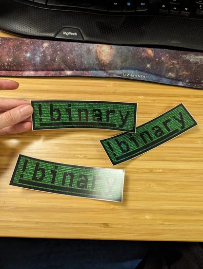 A picture of three identical stickers that say "!binary" with green code-like graphics in the background. They are in a wooden countertop.