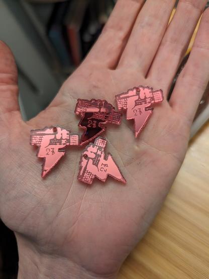 A picture of four shiny pink acrylic charms in the palm of a hand. They look about 1 inch square in size each. They depict a lightning bolt with a fist at the top gripping a keyboard.