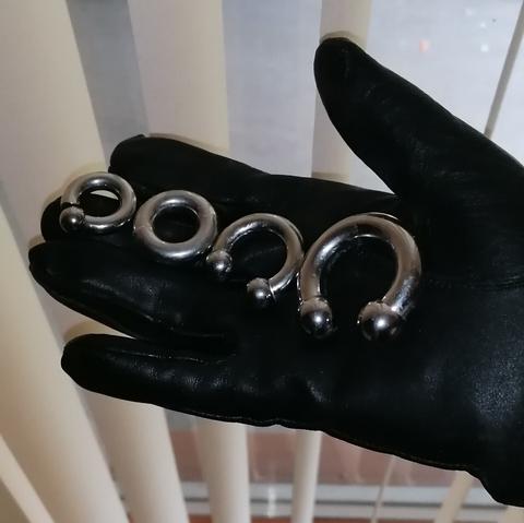 Black leather gloved hand with multiple options of sizes of titanium steel prince Albert piercings to size 00ga.