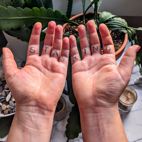 Photo of my hands in front of some houseplants. My palms are open facing the camera, and across the first part of each finger from the palm is "STAY KIND"