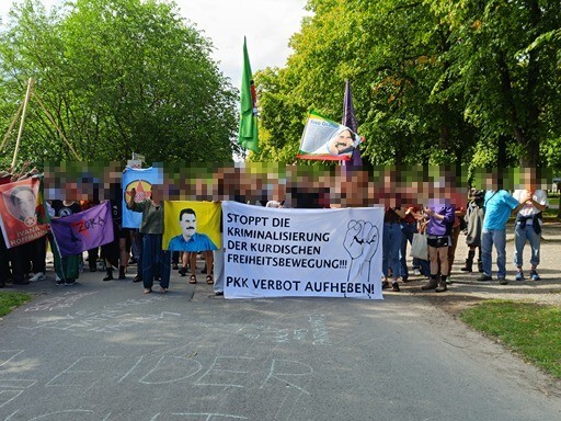 Foto des Protests des internationalistischen Zelts und solidarischen Menschen. Zu sehen sind vielleicht 40 Menschen mit Fahnen und Bannern, die Gesichter der Leute sind verpixelt