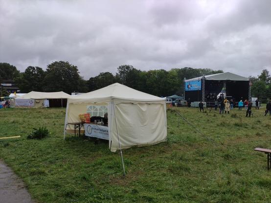 Freifunkstand mit Bühne im rechten Hintergrund auf der eine Band spielt. Ein paar Zuschauer stehen davor. Es regnet.