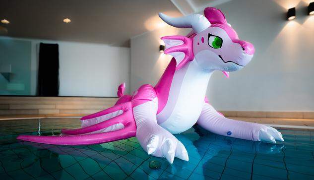 A pink dragon named Ceirsey is floating on a indoor pool. The water has a clear blue tint and it looks like she's shining bright.