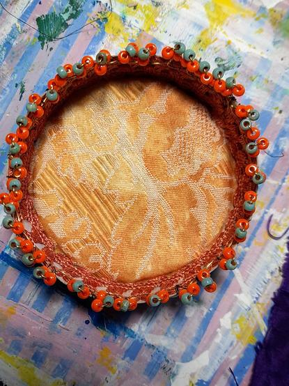 Floral elements of 3 rocailles (in lustred orange and Picasso turquoise) on the finest brass wire are mounted in line at the top of the edges of a round box. The bottom of the box is padded brocade. A reddish-brown braid is lined inside the rim. when everything is stabilised, I have to arrange these elements carefully.