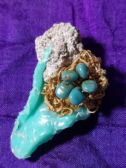 A valuable-looking structure. It is green plastic waste melted and fused with a piece of cement. A kind of flower is mounted in a corner between the two materials. It consists of crocheted golden DCM thread and a stamp made of five drop-shaped glass beads.