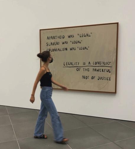 Framed text hanging on a wall (museum?), a girl wearing a (covid) mask walks past, reading the text: 

APARTHEID WAS "LEGAL" 
SLAVERY WAS "LEGAL"
COLONIALISM WAS "LEGAL"

LEGALITY IS A CONSTRUCT OF THE POWERFUL NOT OF JUSTICE