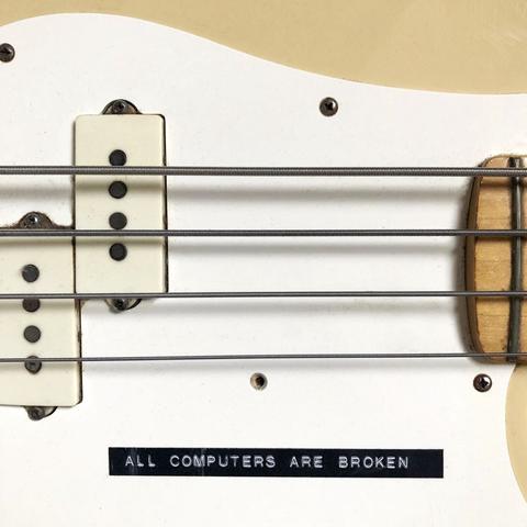 A close-up of a beige electric bass guitar with a white pickguard. Also two cream coloured pickups and four strings. Underneath te strongs is a sticker with the text ALL COMPUTERS ARE BROKEN.