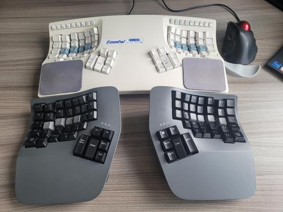 Two keyboards and a trackball on a table. Bottom keyboard is a new Kinesis Advantage360 Professional, top keyboard is a pre-PS/2-era over-30-years-old Kinesis-not-even-Advantage-yet.